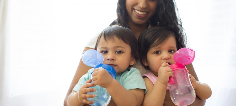 Two babies holding blue and pink cups sitting in mother's lap